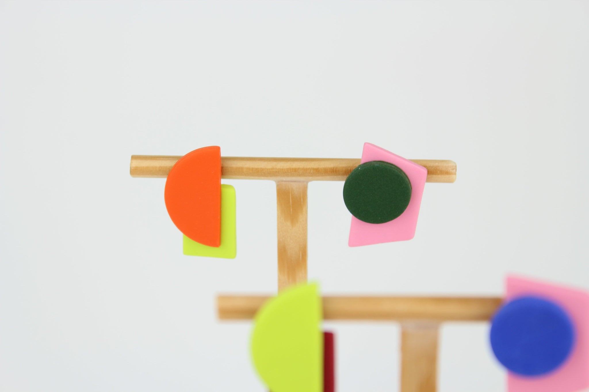 Mehrere asymmetrische Ohrringe aus Polymerton mit geometrischen Formen in kräftigen Farben, arrangiert auf hölzernen Schmuckständern vor hellem Hintergrund.