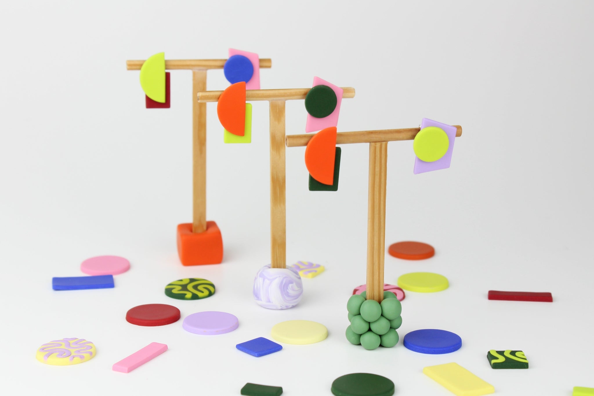 Geometrische Ohrhänger von Atelier Anda auf Schmuckständern arrangiert, umgeben von farbigen Polymerton-Elementen in verschiedenen Formen.