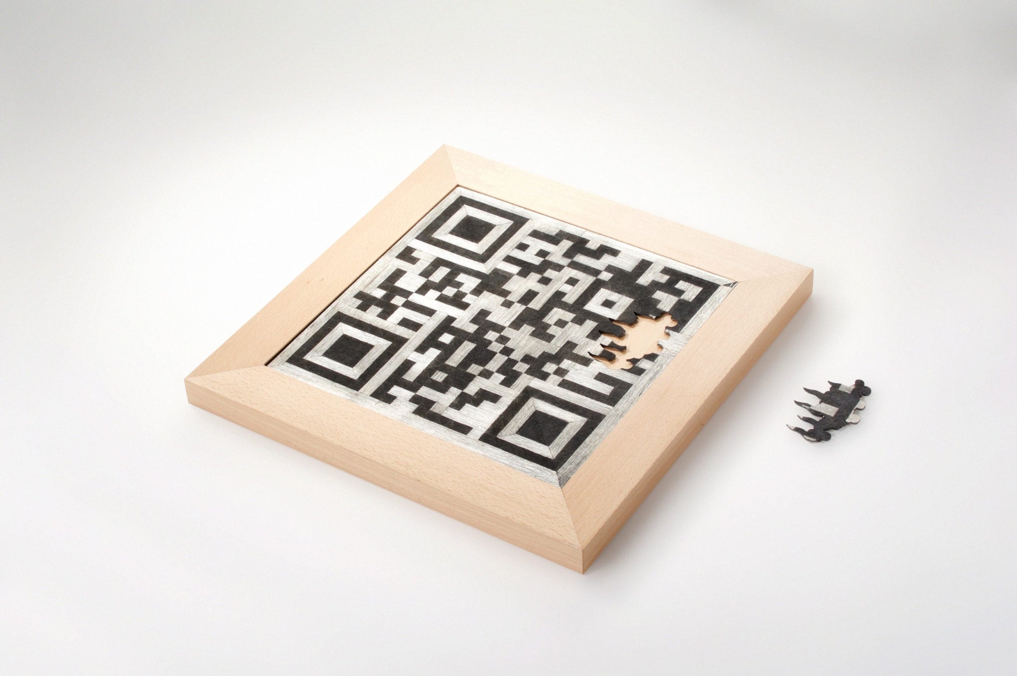 Ein in Holz gerahmter QR-Code, der aus  schwarzem Eisen und Silberdraht-Intarsien besteht, aus dem Code kann eine kleine Brosche entnommen werdne. 