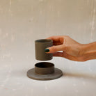 Modularer Espressobecher mit Untertasse aus der Kollektion ROSA MORADA von Martina Geroni in der Tonfarbe Buccia di Zenzero, gezeigt im Moment des Abhebens des Bechers von der flachen, zylindrischen Untertasse aus handgefertigtem Steingut.