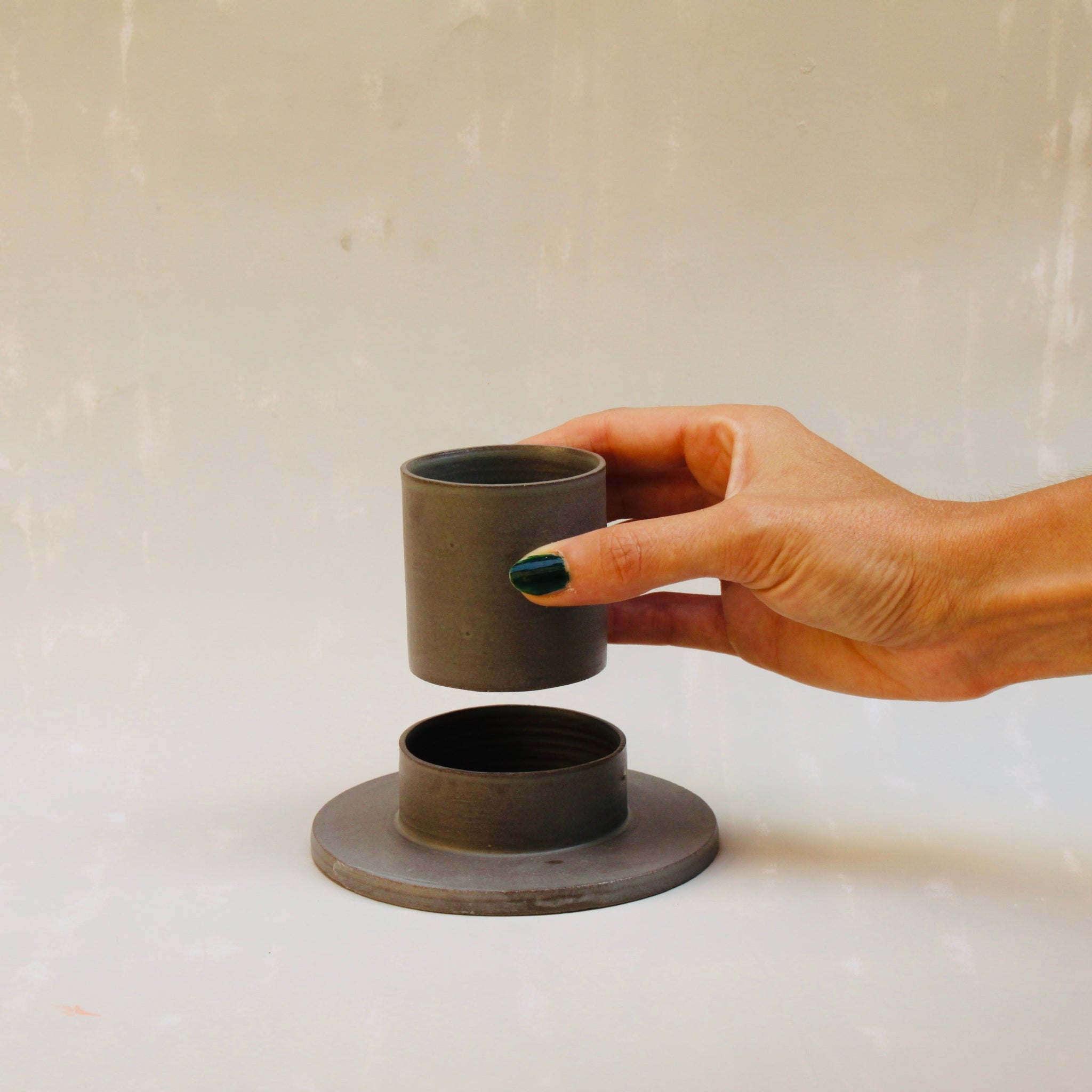 Eine Hand hält einen modularen Espressobecher mit Untertasse aus rauchgrauem Steingut der Genueser Künstlerin Martina Geroni.