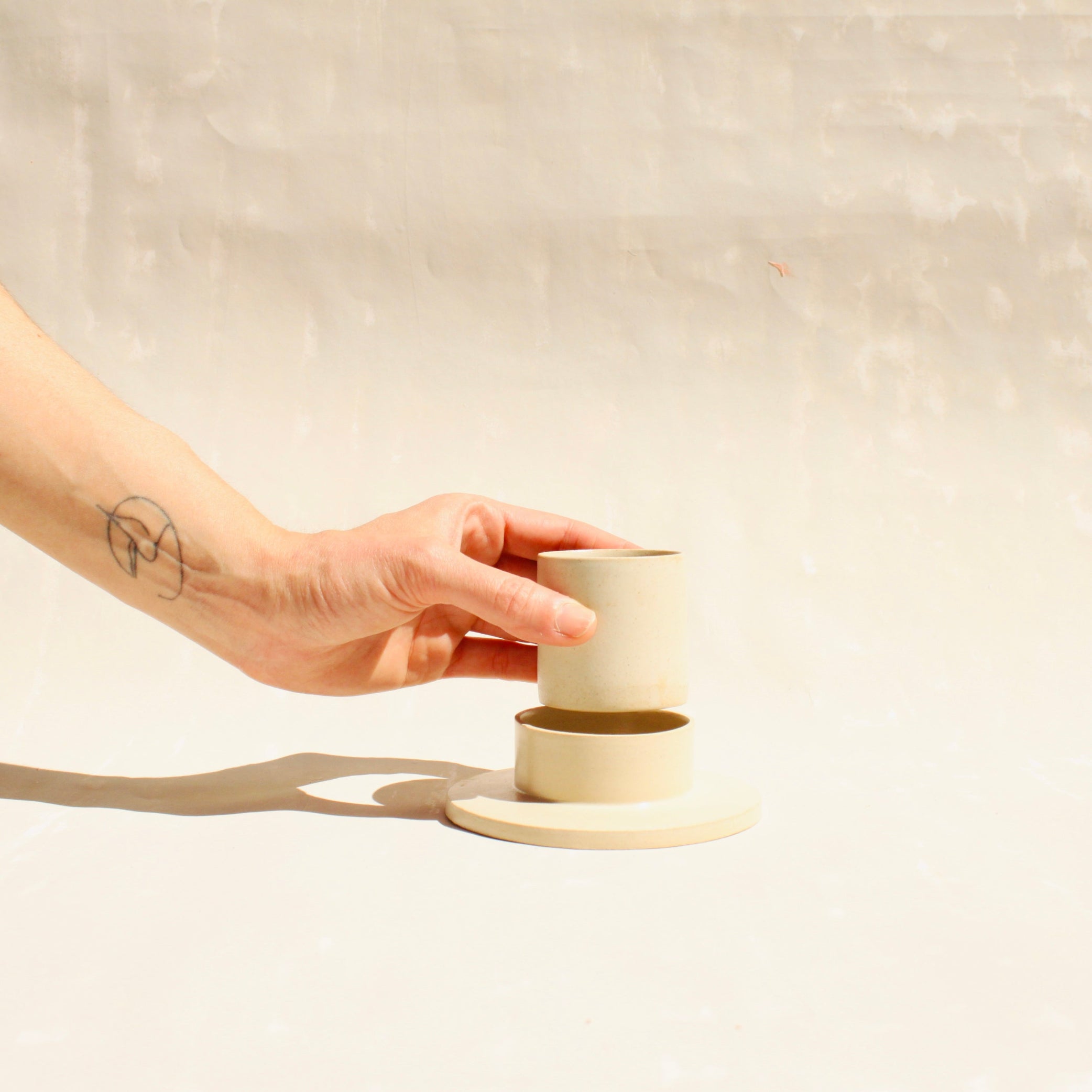 Modularer Espressobecher mit Untertasse aus der Kollektion ROSA MORADA von Martina Geroni in der Tonfarbe Fiore di Cotone, handgefertigt aus Steingut, gezeigt beim Abheben des Bechers vom Unterschälchen.