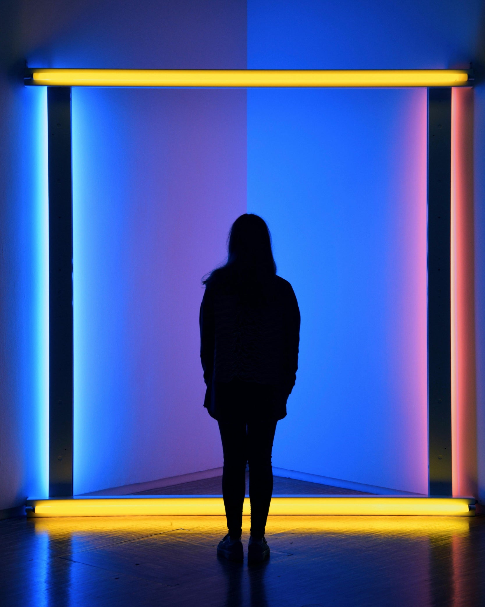 Die Schattensilhouette einer Frau steht vor einer Kunstinstallation von Dan Flavin mit farbigen Neonröhren, die in der Ecken eines Raum diagonal aufgebaut sind, die Szenerie leuchtet dunkleblau.