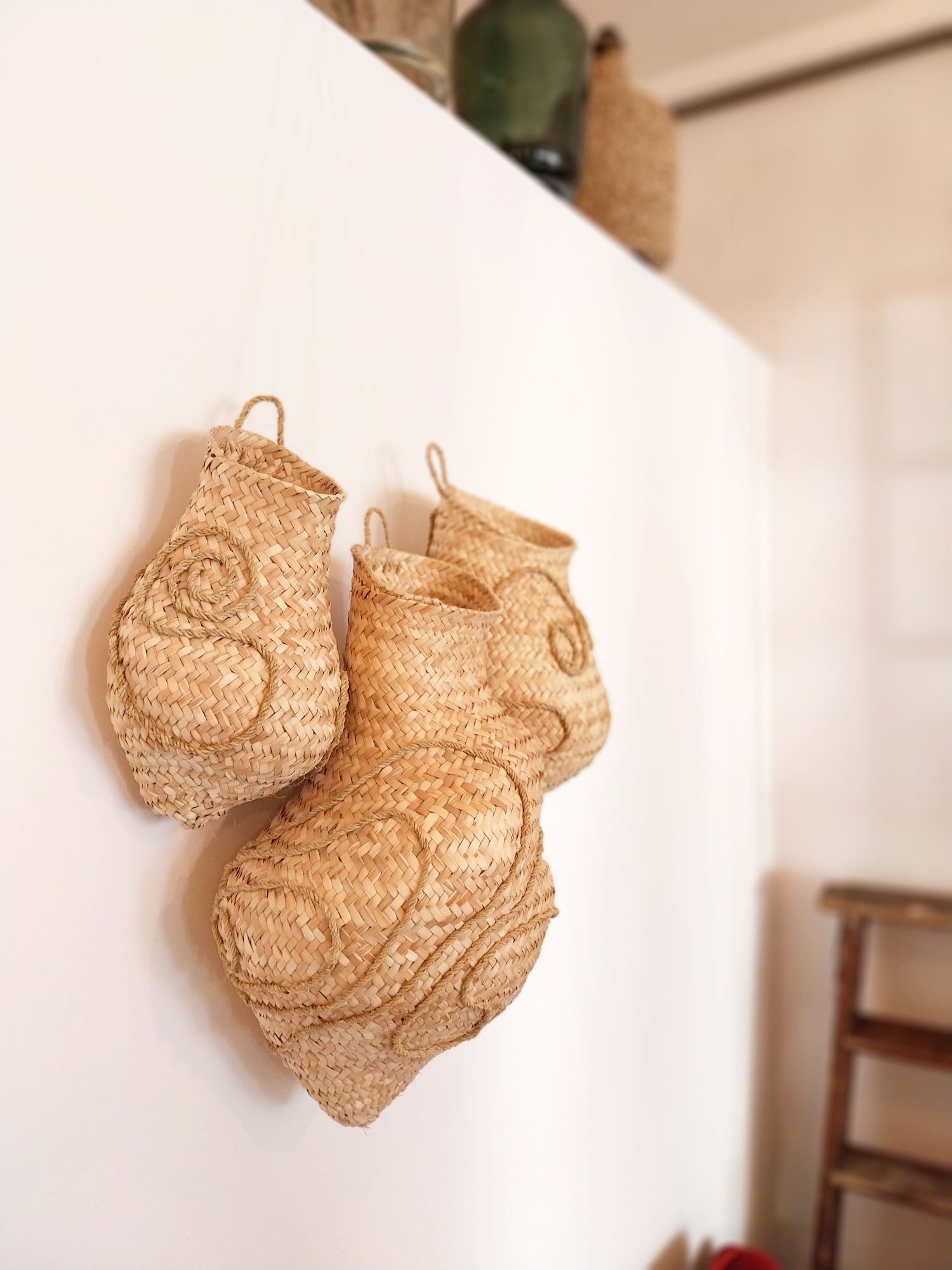 Wandskulptur aus drei handgeflochtenen Körben aus hellen Palmwedeln, organisch geformt und mit reliefartigen Spiral- und Linienmustern versehen, dicht nebeneinander an einer weißen Wand montiert; die natürliche Faserstruktur und warmen Beigetöne stehen im Kontrast zum ruhigen, minimalistischen Raum.