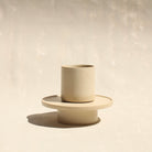 Modularer Mokkabecher mit Untertasse aus der Kollektion ROSA MORADA von Martina Geroni in der Tonfarbe Fiore di Cotone, handgefertigt aus Steingut, der Becher auf dem Unterschälchen als Podest stehend.