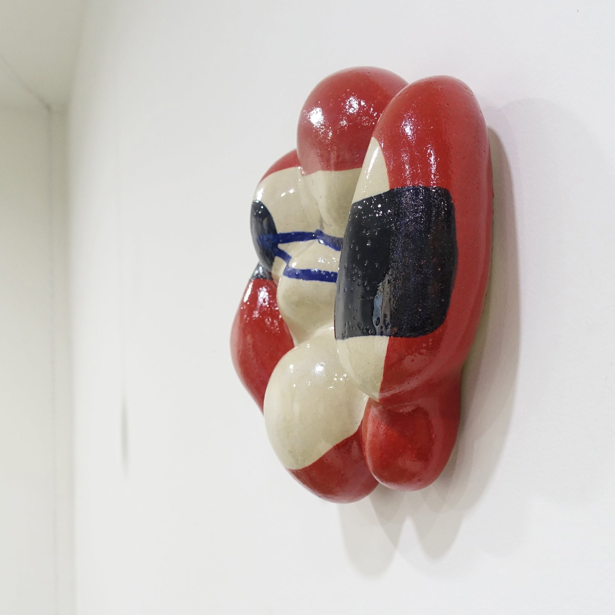 Seitenansicht einer Wandskulptur aus Keramik mit organisch gewölbter Form; cremefarbener Grundton mit leuchtend roten Flächen sowie dunkelblauen, gestischen Linien- und Flächenakzenten auf der Oberfläche.