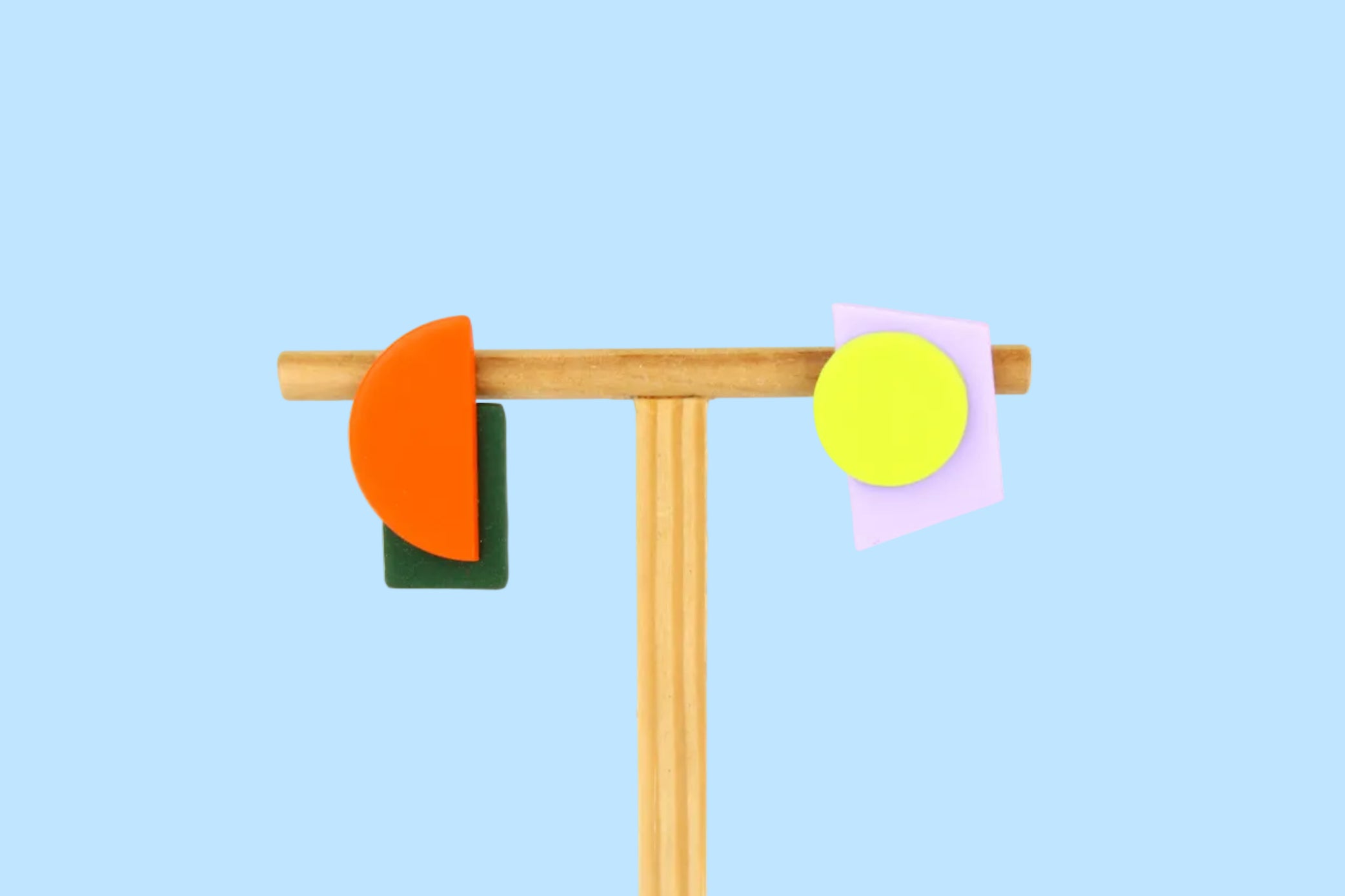 Asymmetrische Ohrringe aus Polymerton mit grafischen Formen in Orange, Dunkelgrün, Neongelb und Flieder, präsentiert auf einem schlichten Schmuckständer vor pastellfarbenem Hintergrund.