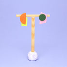 Asymmetrische Ohrringe aus Polymerton mit grafischen Formen in Orange, Grün, Gelb und Rosa, präsentiert auf einem schlichten Schmuckständer vor pastellfarbenem Hintergrund.