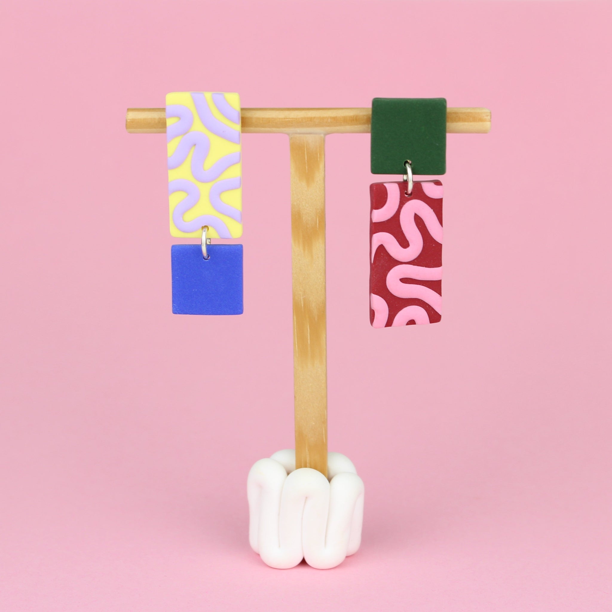 Asymmetrische Ohrringe aus Polymerton mit rechteckigen Elementen und grafischen Mustern in Gelb, Rot, Flieder, Blau und Grün, präsentiert auf einem hölzernen Schmuckständer.