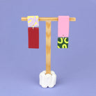 Asymmetrische Ohrringe aus Polymerton mit rechteckigen Elementen und grafischen Mustern in Rot, Rosa, Buttergelb, Grün und Flieder, präsentiert auf einem hölzernen Schmuckständer.