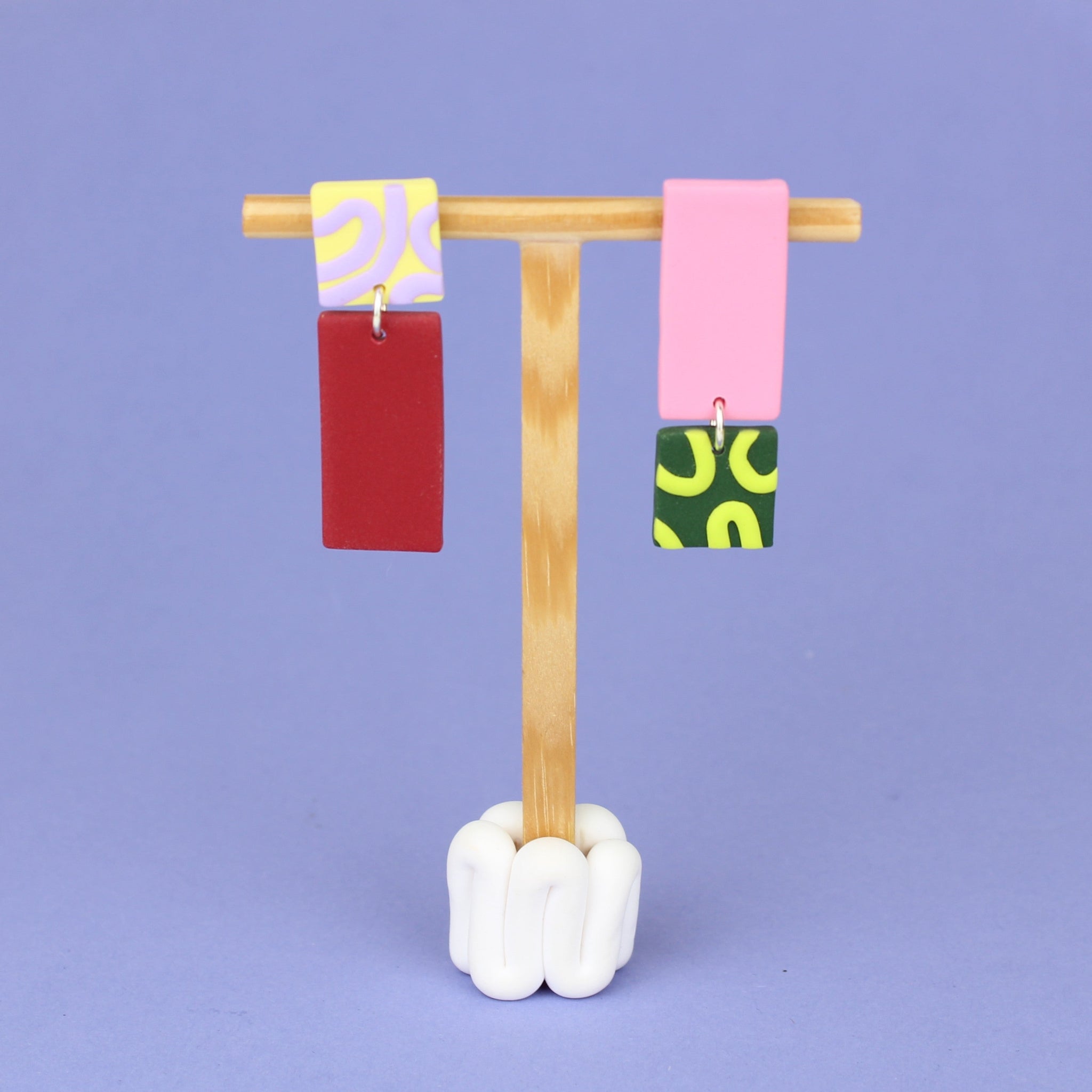 Asymmetrische Ohrringe aus Polymerton mit rechteckigen Elementen und grafischen Mustern in Rot, Rosa, Buttergelb, Grün und Flieder, präsentiert auf einem hölzernen Schmuckständer.