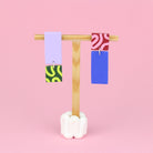 Asymmetrische Ohrringe aus Polymerton mit rechteckigen Elementen und grafischen Mustern in Rosa und Grün, präsentiert auf einem hölzernen Schmuckständer vor rosa Hintergrund.