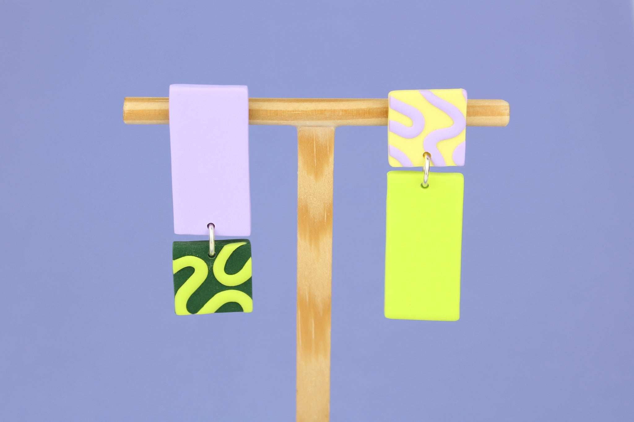 Asymmetrische Ohrringe aus Polymerton mit rechteckigen Elementen und grafischen Mustern in Flied, präsentiert auf einem hölzernen Schmuckständer.