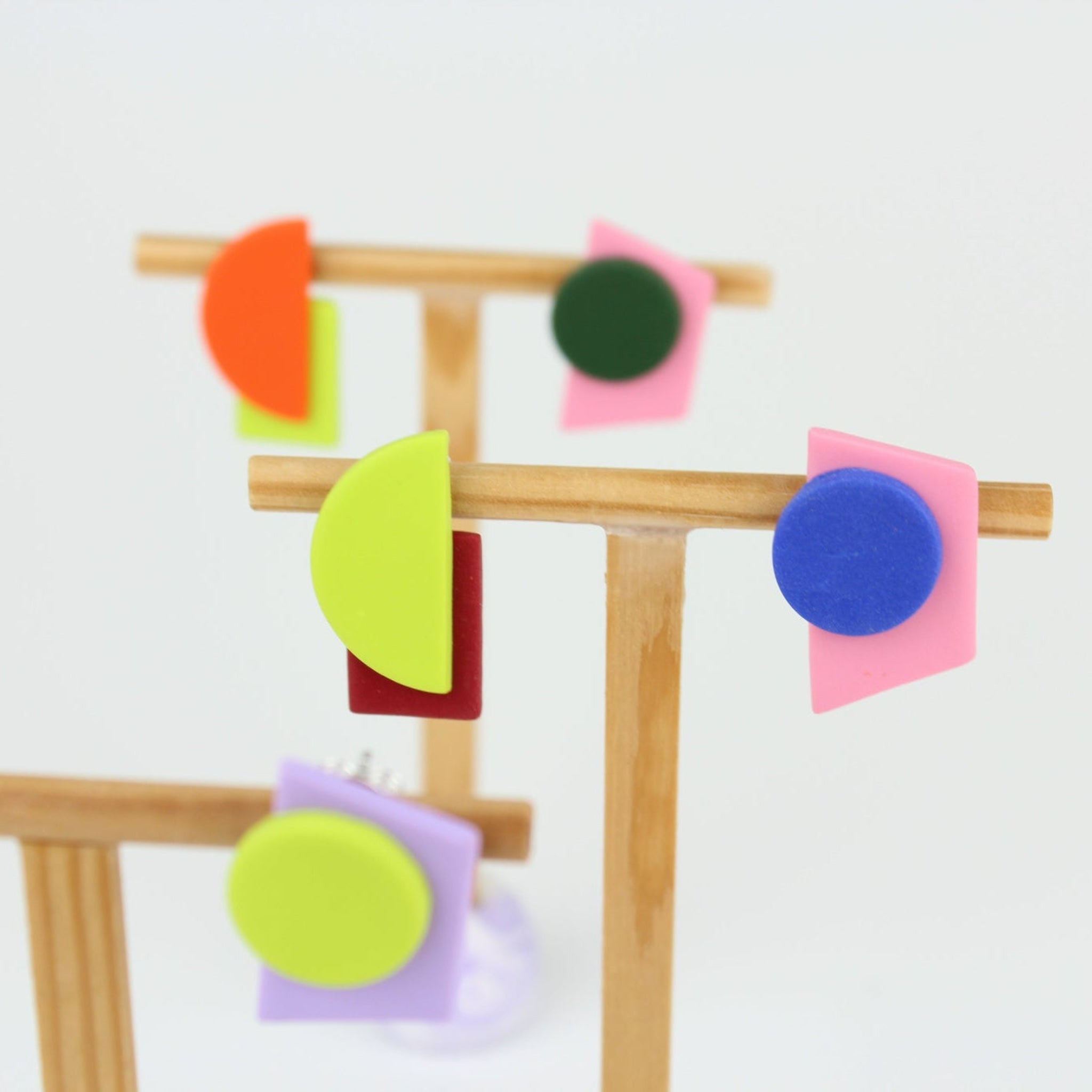 Mehrere asymmetrische Ohrringe aus Polymerton mit geometrischen Formen in kräftigen Farben, arrangiert auf hölzernen Schmuckständern vor hellem Hintergrund.