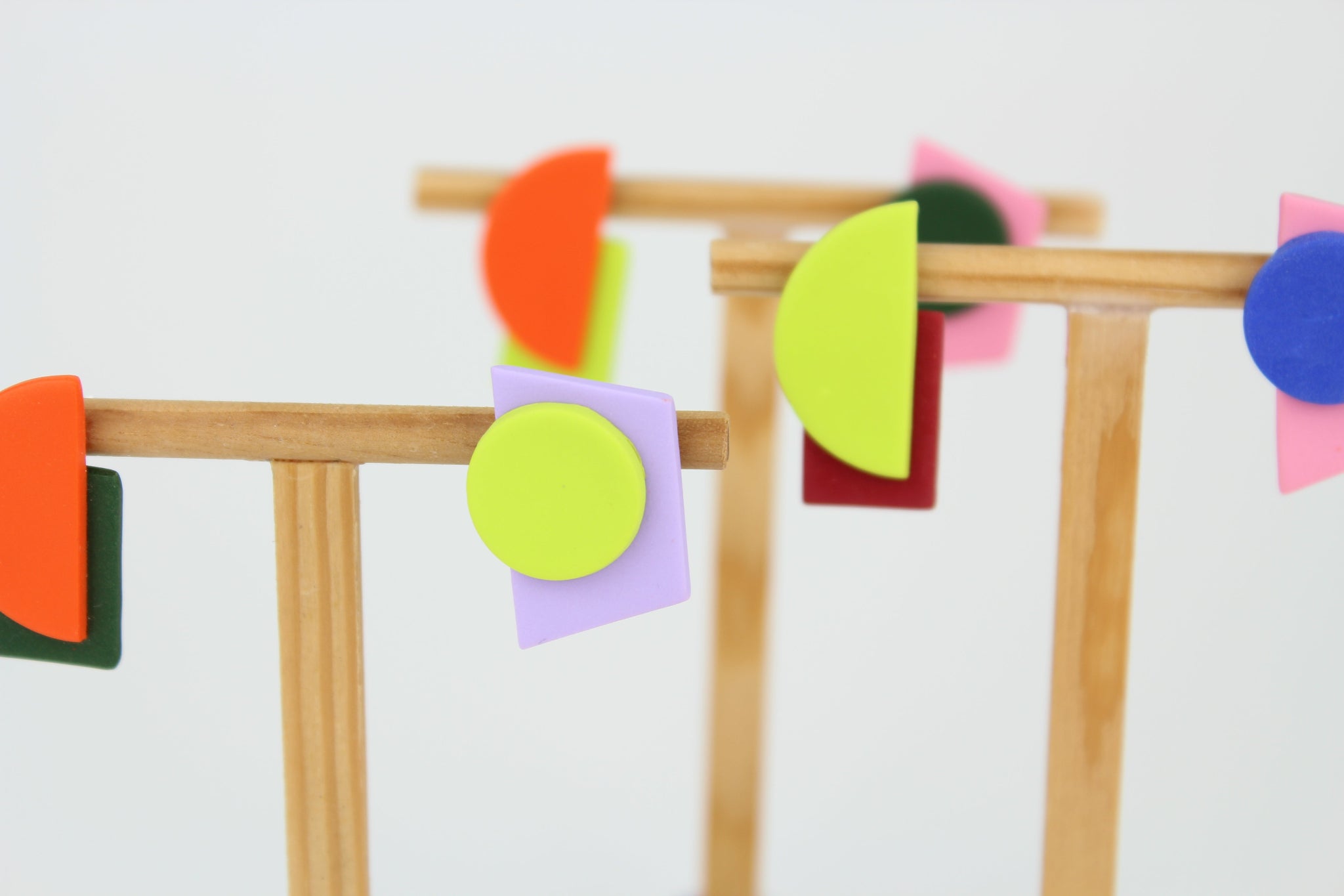 Mehrere asymmetrische Ohrringe aus Polymerton mit geometrischen Formen in kräftigen Farben, arrangiert auf hölzernen Schmuckständern vor hellem Hintergrund.