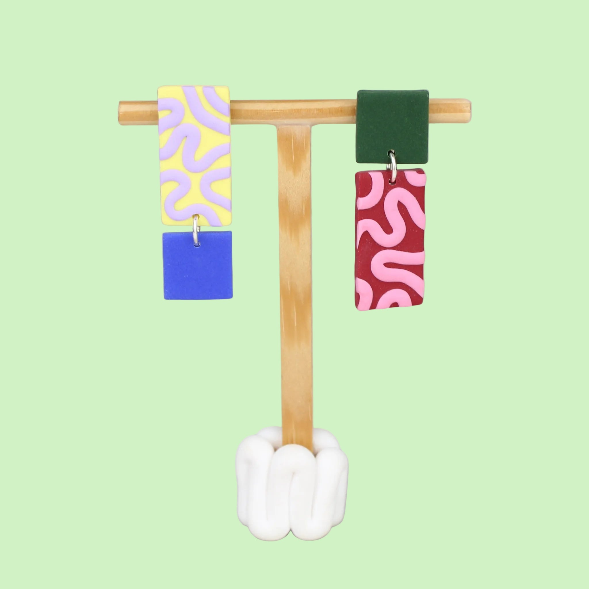 Asymmetrische Ohrringe aus Polymerton mit rechteckigen Elementen und grafischen Mustern in Gelb, Rot, Flieder, Blau und Grün, präsentiert auf einem hölzernen Schmuckständer.