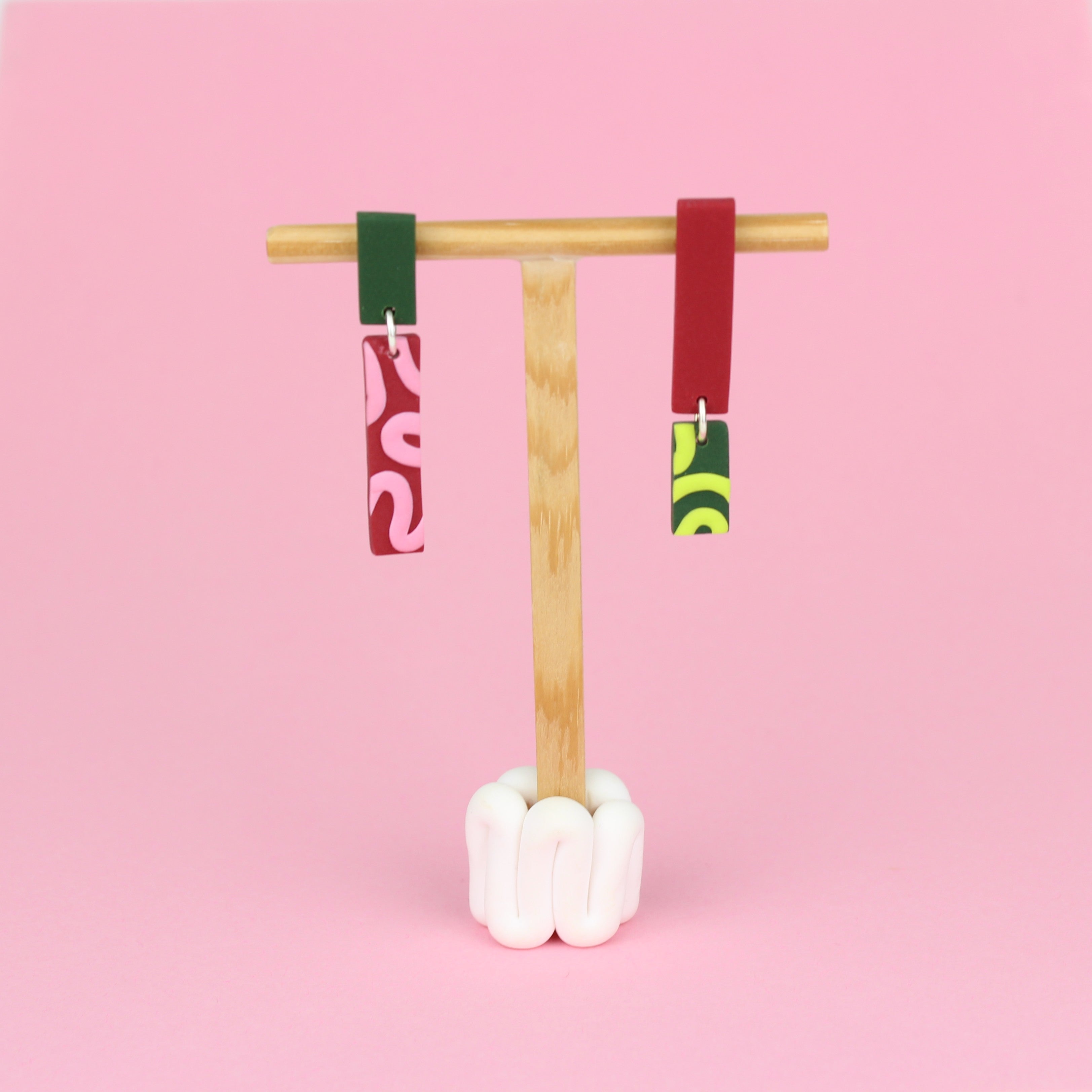 Asymmetrische Ohrringe aus Polymerton mit schmalen rechteckigen Elementen und grafischen Mustern in Rot, Rosa, Grün und Neongelb, präsentiert auf einem hölzernen Schmuckständer.