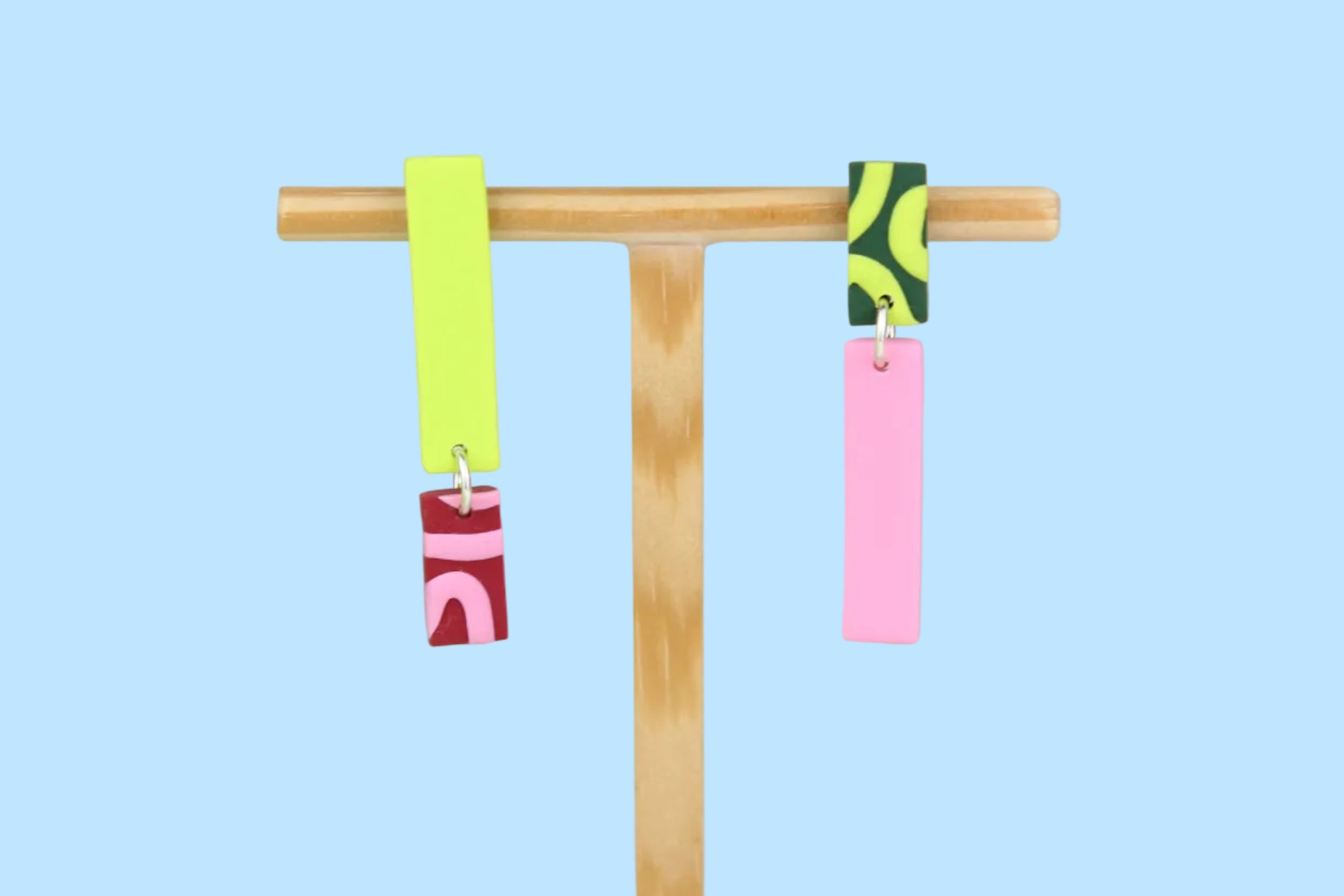 Asymmetrische Ohrringe aus Polymerton mit schmalen rechteckigen Elementen und grafischen Mustern in Rot, Rosa, Grün und Neongelb, präsentiert auf einem hölzernen Schmuckständer.