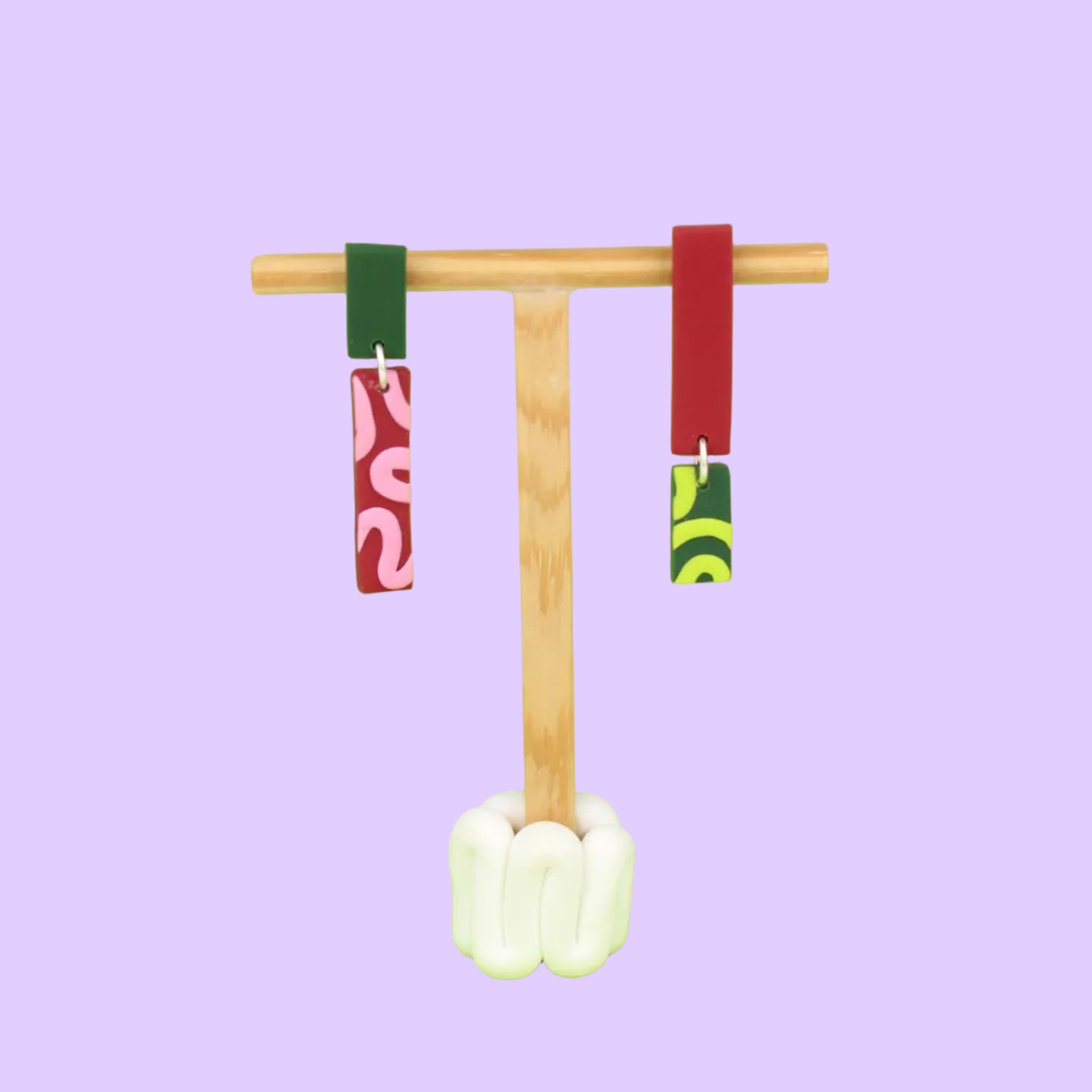Asymmetrische Ohrringe aus Polymerton mit schmalen rechteckigen Elementen und grafischen Mustern in Rot, Rosa, Grün und Neongelb, präsentiert auf einem hölzernen Schmuckständer.