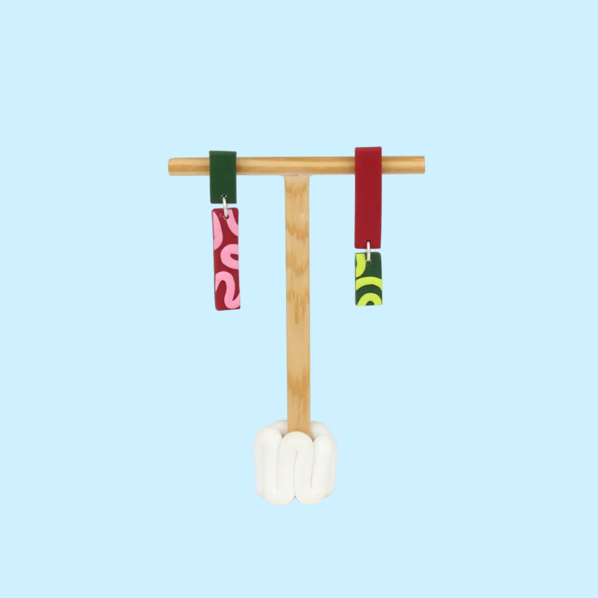 Asymmetrische Ohrringe aus Polymerton mit schmalen rechteckigen Elementen und grafischen Mustern in Rot, Rosa, Grün und Neongelb, präsentiert auf einem hölzernen Schmuckständer.