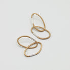 Ein Paar Ohrringe aus je zwei ovalen Gliedern mit charakteristischer Haargummistruktur und Bördelungsdetail an der Schliesse, minimalistisch und handgefertigt. Vergoldetes Silber mit silberner Schliesse, arrangiert auf hellem Hintergrund.