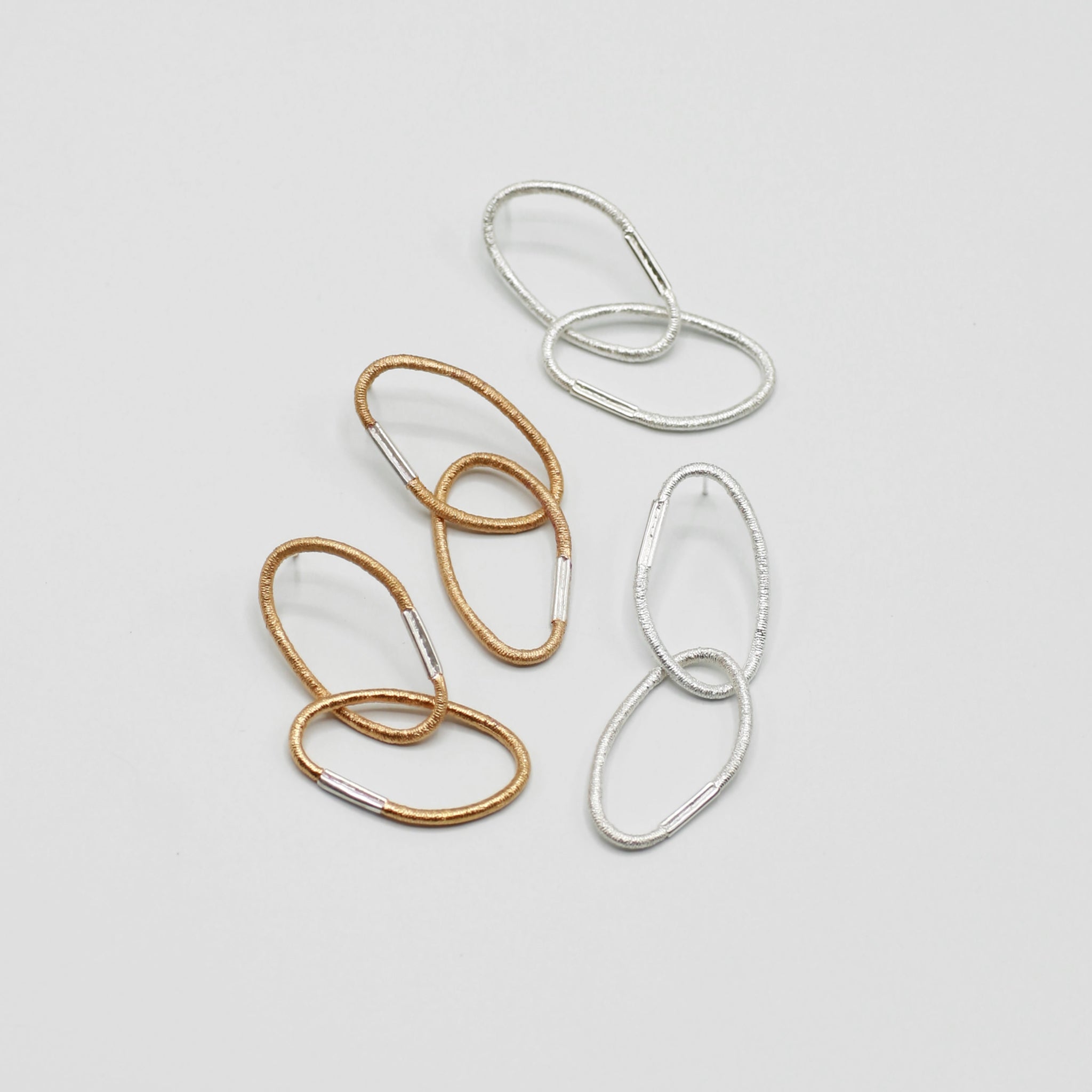 Zwei Paar Ohrringe aus je zwei ovalen Gliedern mit charakteristischer Haargummistruktur und Bördelungsdetail an der Schliesse, minimalistisch und handgefertigt. Ein Paar Vermeil mit silberner Schliesse, ein Paar Silber mit silberner Schliesse, arrangiert auf hellem Hintergrund.