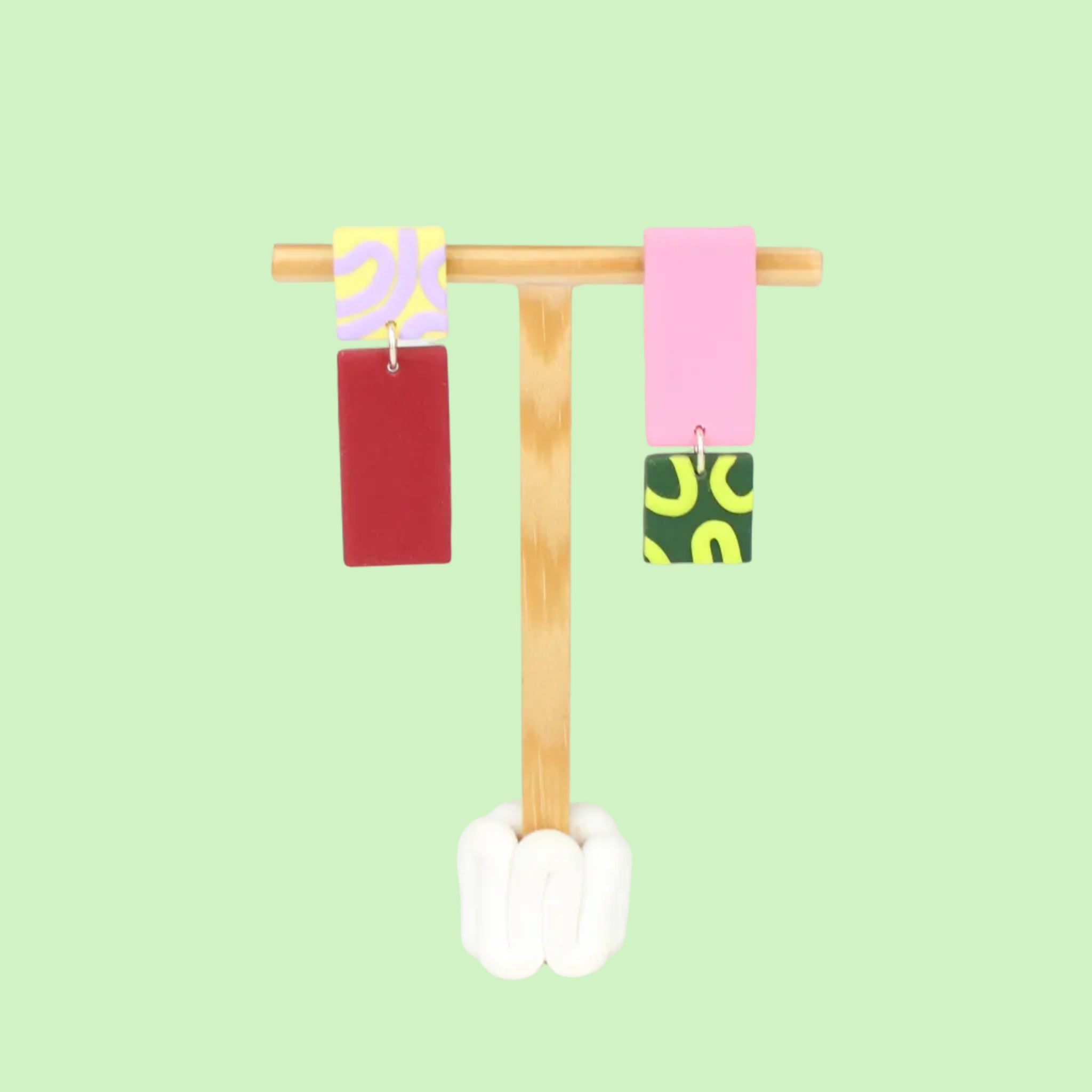 Asymmetrische Ohrringe aus Polymerton mit rechteckigen Elementen und grafischen Mustern in Flied, präsentiert auf einem hölzernen Schmuckständer.