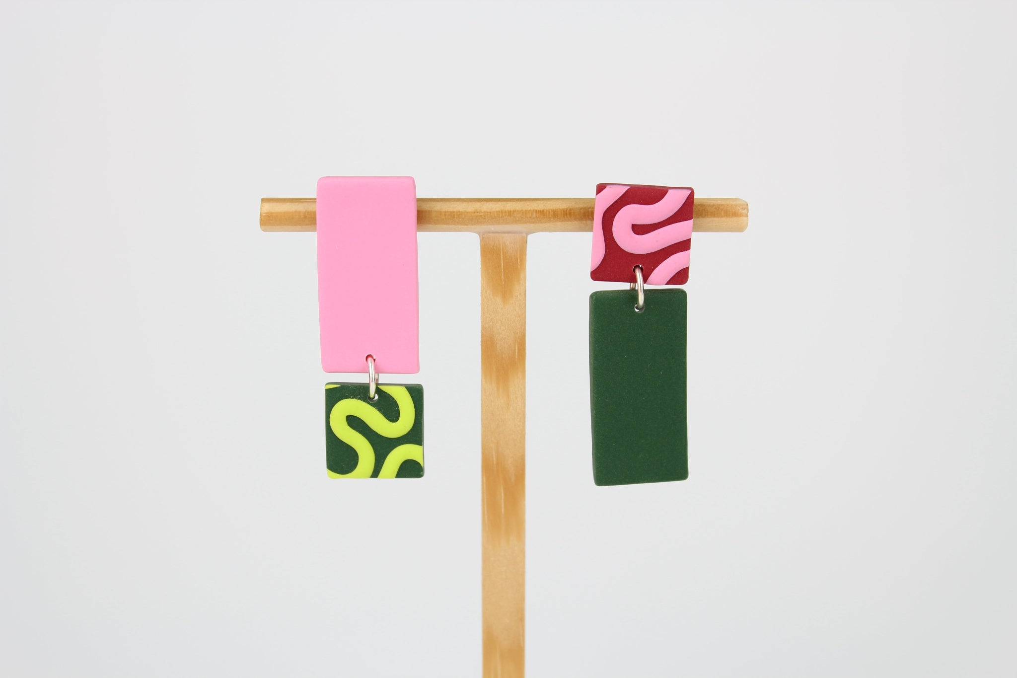 Asymmetrische Ohrringe aus Polymerton mit rechteckigen Elementen und grafischen Mustern in Rosa und Grün, präsentiert auf einem hölzernen Schmuckständer vor hellgrünem Hintergrund.