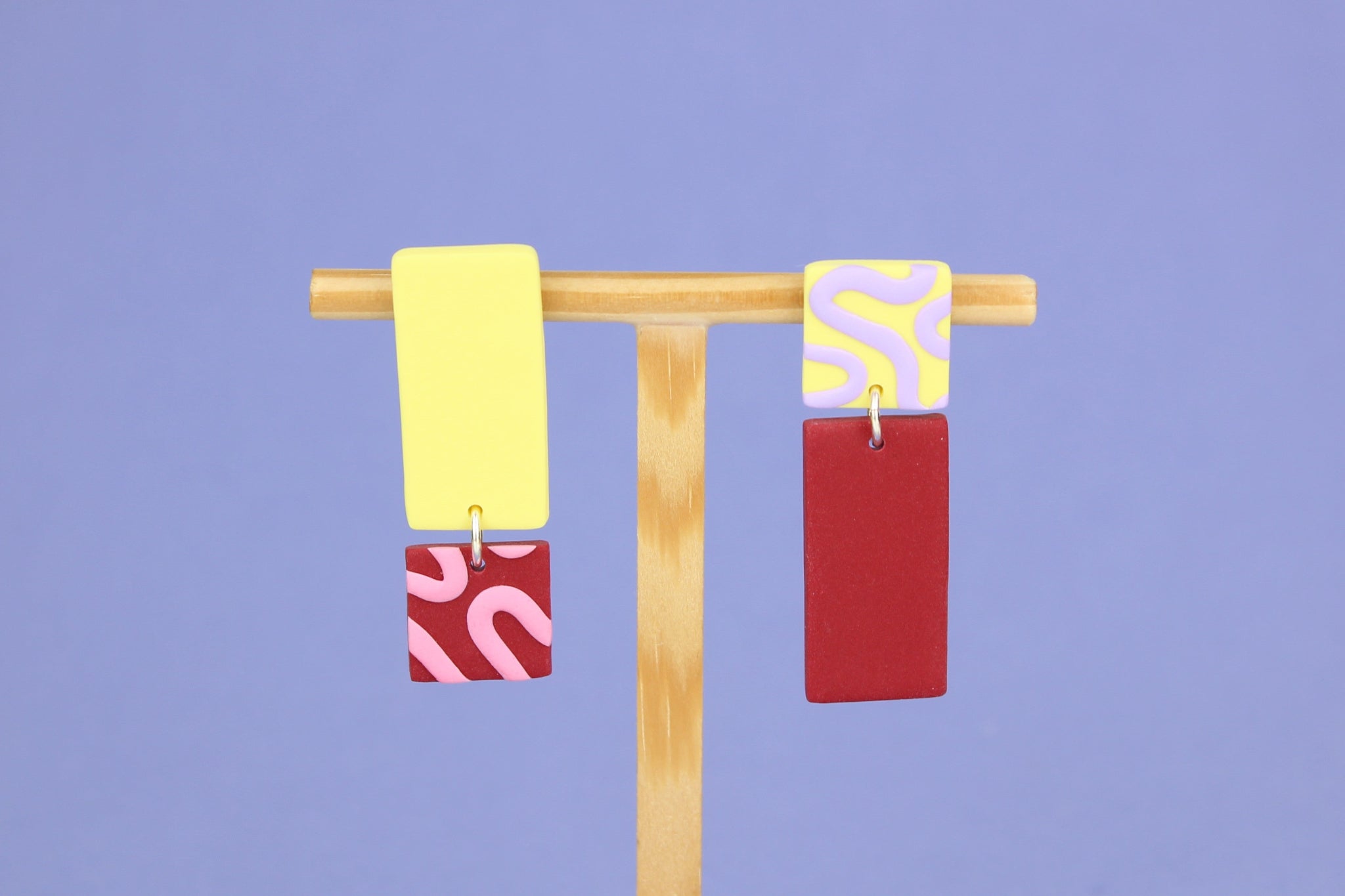 Asymmetrische Ohrringe aus Polymerton mit rechteckigen Elementen und grafischen Mustern in Gelb, Rot, Rosa und Flieder, präsentiert auf einem hölzernen Schmuckständer.