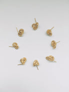 Acht kleine goldene Ohrstecker in unterschiedlichen Knotenform liegen in einem Kreis auf einem weissen Studiohintergrund. 