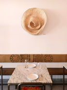 Wandskulptur PALMA V aus handgeflochtenen Palmwedeln, organisch geformt und schalenartig vertieft, ähnelt einem riesigen zerdrückten Strohhut, an einer hellen Wand über einem gedeckten Marmortisch montiert.