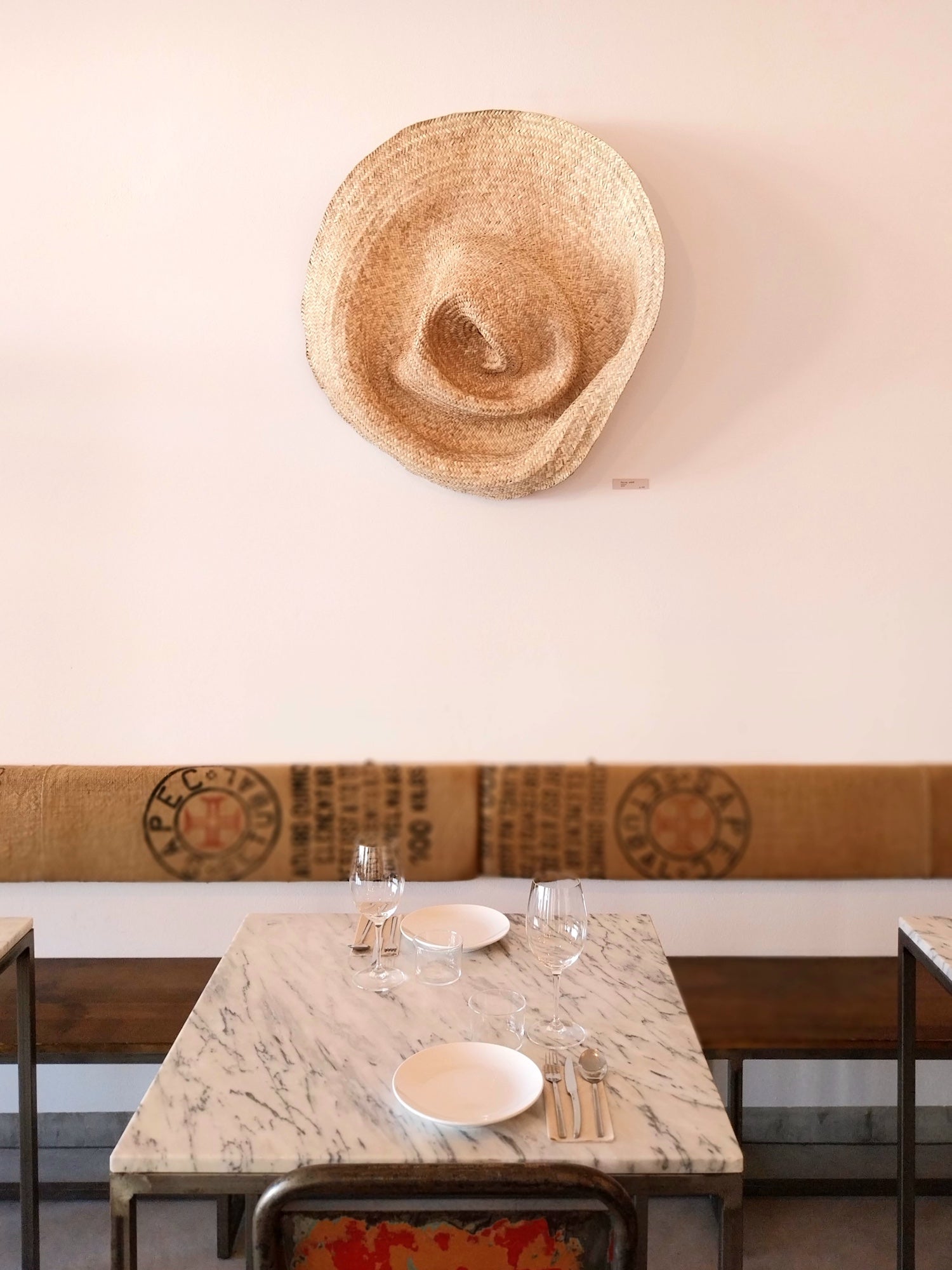 Wandskulptur PALMA V aus handgeflochtenen Palmwedeln, organisch geformt und schalenartig vertieft, ähnelt einem riesigen zerdrückten Strohhut, an einer hellen Wand über einem gedeckten Marmortisch montiert.