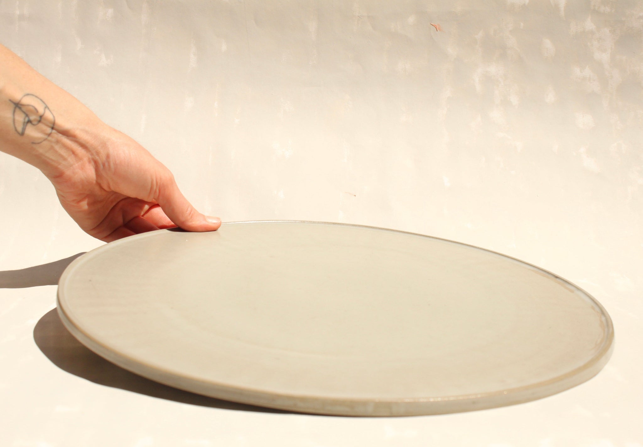 Modularer Pizzateller aus der Kollektion ROSA MORADA von Martina Geroni in der Tonfarbe Fiore di Cotone aus Steingut, flach ausgeführt und von Hand gehalten, nutzbar als Pizzateller oder als reduziertes Platzset.