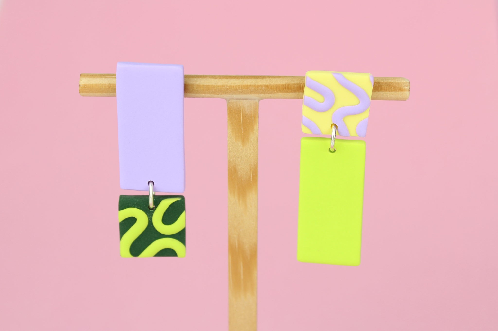 Asymmetrische Ohrringe aus Polymerton mit rechteckigen Elementen und grafischen Mustern in Flied, präsentiert auf einem hölzernen Schmuckständer.