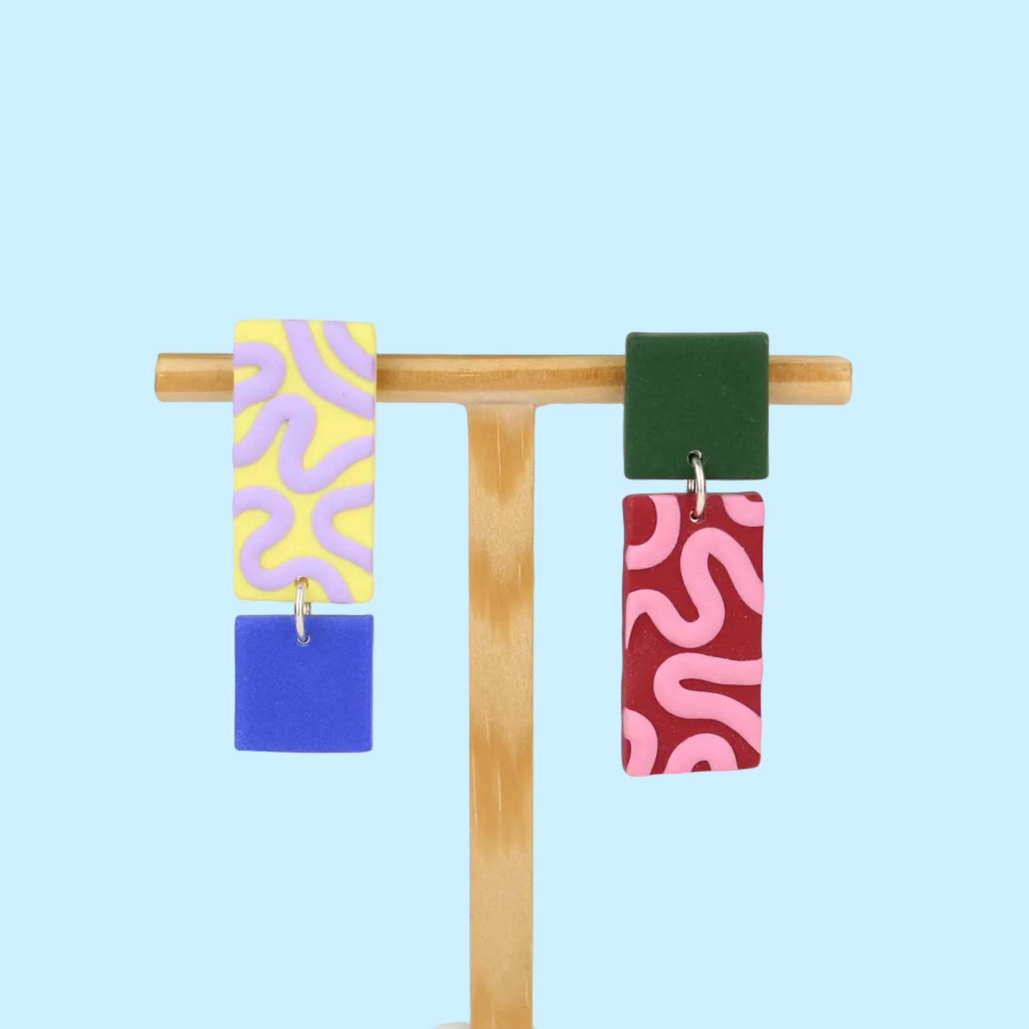 Asymmetrische Ohrringe aus Polymerton mit rechteckigen Elementen und grafischen Mustern in Gelb, Rot, Flieder, Blau und Grün, präsentiert auf einem hölzernen Schmuckständer.