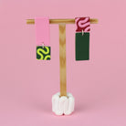 Asymmetrische Ohrringe aus Polymerton mit rechteckigen Elementen und grafischen Mustern in Rosa und Grün, präsentiert auf einem hölzernen Schmuckständer vor hellgrünem Hintergrund.