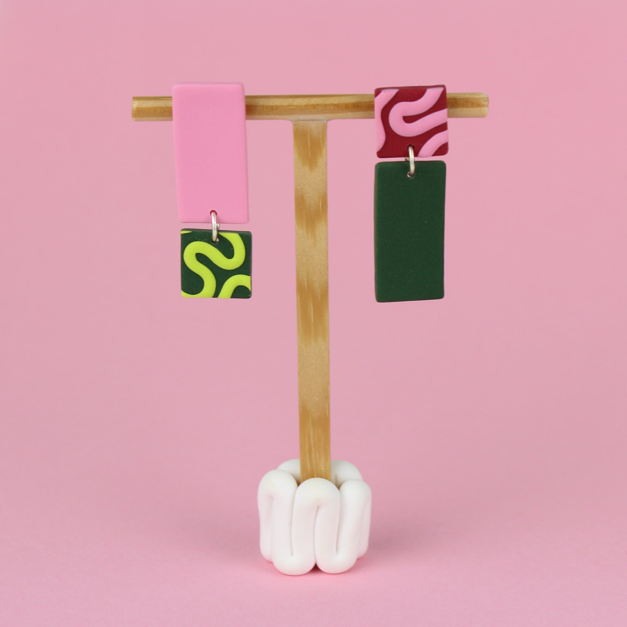 Asymmetrische Ohrringe aus Polymerton mit rechteckigen Elementen und grafischen Mustern in Rosa und Grün, präsentiert auf einem hölzernen Schmuckständer vor hellgrünem Hintergrund.