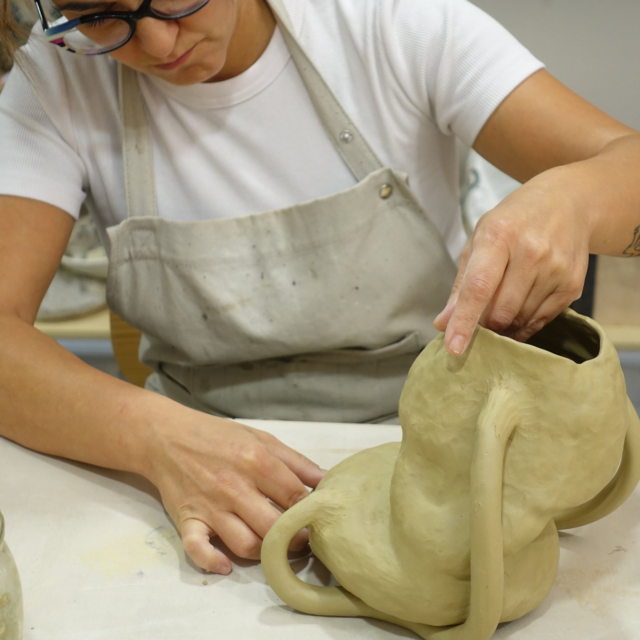 Die Keramikerin Alice del Ferraro formt an einem Arbeitstisch ein organisch geschwungenes Gefäß aus hellem Ton. Sie trägt eine hellgraue Schürze und arbeitet konzentriert mit beiden Händen am noch weichen Objekt.