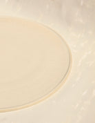 Detailansicht der flachen  Tischsetzs aus der Kollektion ROSA MORADA von Martina Geroni in der Tonfarbe Fiore di Cotone, gefertigt aus Steingut, mit minimal erhöhtem Rand auf hellem Hintergrund.