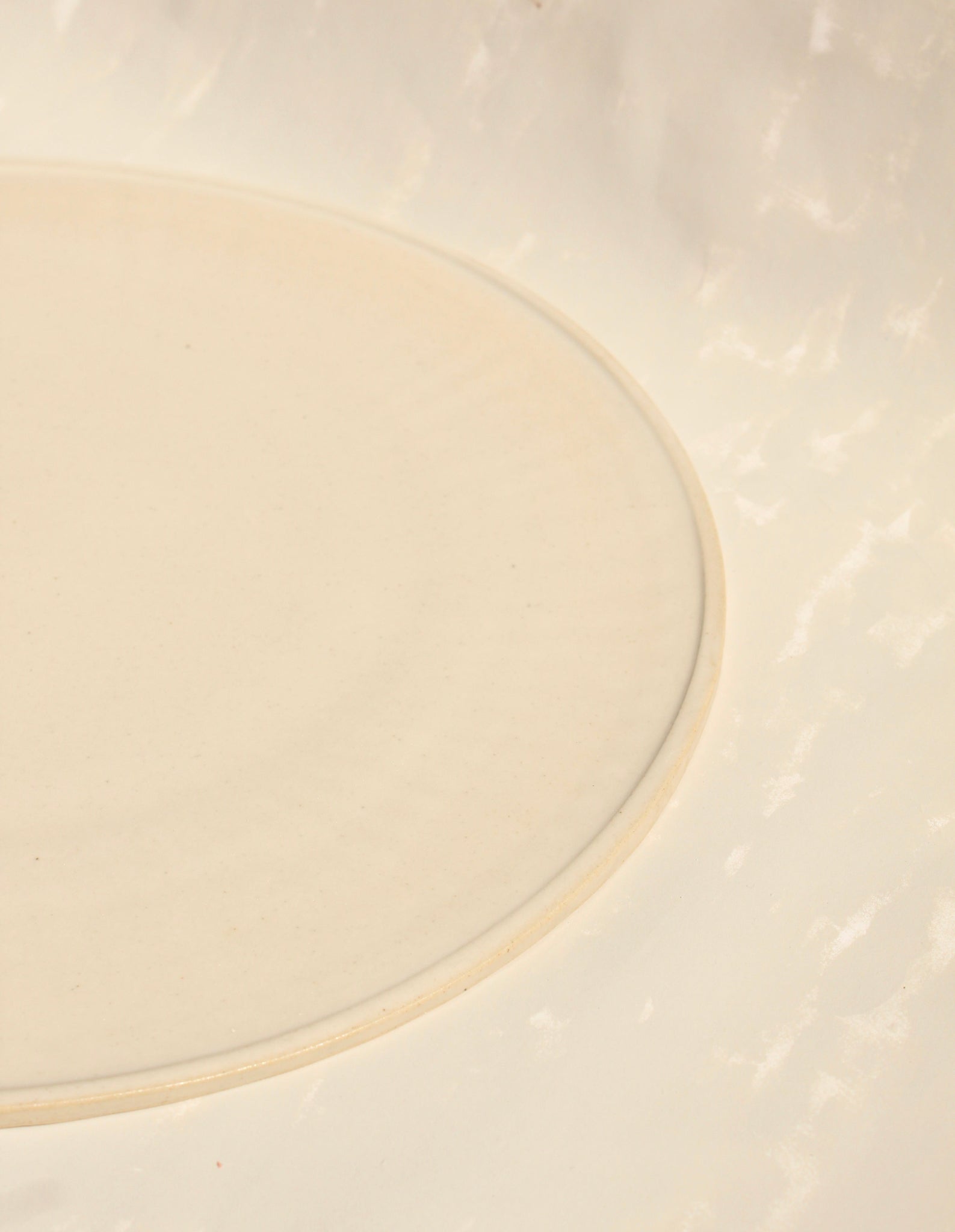 Detailansicht der flachen  Tischsetzs aus der Kollektion ROSA MORADA von Martina Geroni in der Tonfarbe Fiore di Cotone, gefertigt aus Steingut, mit minimal erhöhtem Rand auf hellem Hintergrund.