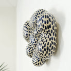 Seitenansicht einer Wandskulptur aus Keramik mit organisch gewölbter, wolkenähnlicher Form; helles Grundmaterial, dicht überzogen mit gleichmäßig verteilten dunkelblauen, tropfenförmigen Pinselstrichen.