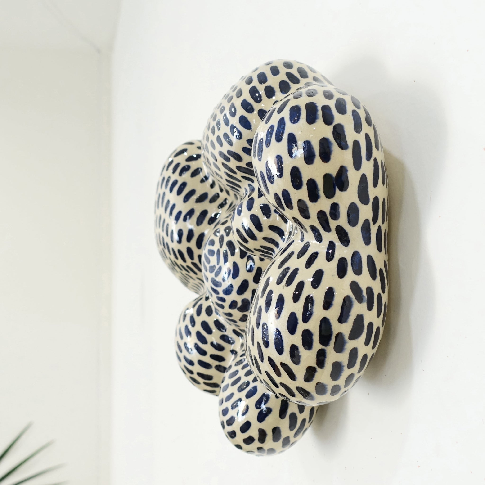 Seitenansicht einer Wandskulptur aus Keramik mit organisch gewölbter, wolkenähnlicher Form; helles Grundmaterial, dicht überzogen mit gleichmäßig verteilten dunkelblauen, tropfenförmigen Pinselstrichen.