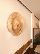 Schräge Seitenansicht der Wandskulptur PALMA V aus handgeflochtenen Palmwedeln, organisch geformt und schalenartig vertieft, ähnelt einem riesigen zerdrückten Strohhut, an einer hellen Wand in einem Galerieartigen Raum mit sitzbänken und Tischchen. 