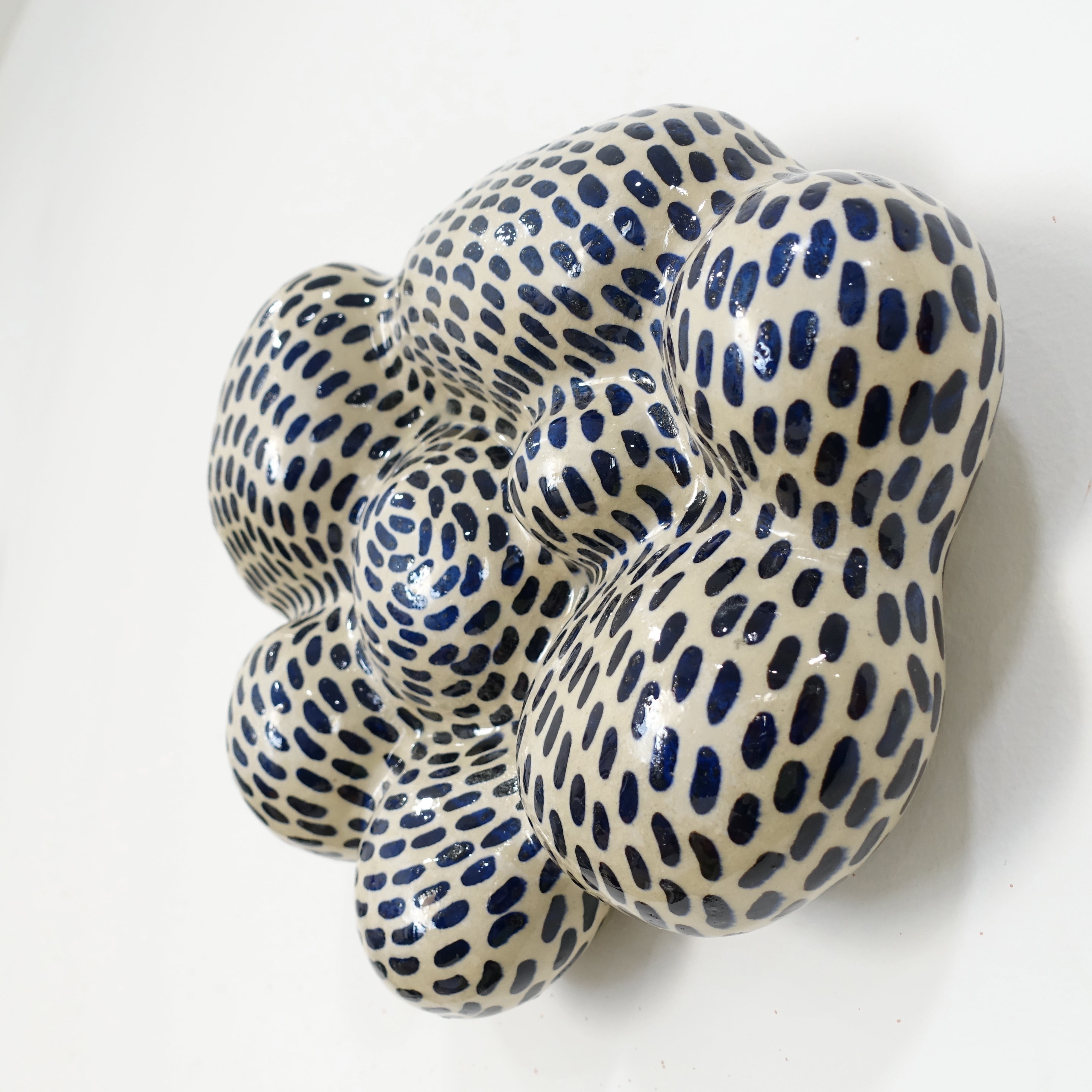 Seitenansicht einer keramischen Wandskulptur mit organisch gewölbter, wolkenähnlicher Form; helles Grundmaterial, dicht überzogen mit gleichmäßig verteilten dunkelblauen, tropfenförmigen Pinselstrichen.