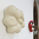 Seitenansicht einer keramischen Wandskulptur in organischer, wolkenartiger Form; helle, leicht cremeweiße Oberfläche mit feinen Rissstrukturen und weicher, gewölbter Modellierung. Im Hintergrund die Nuvola Rossa mit hellem Grundton, roten Flächen und abstrakten schwarzen und blauen Mustern.