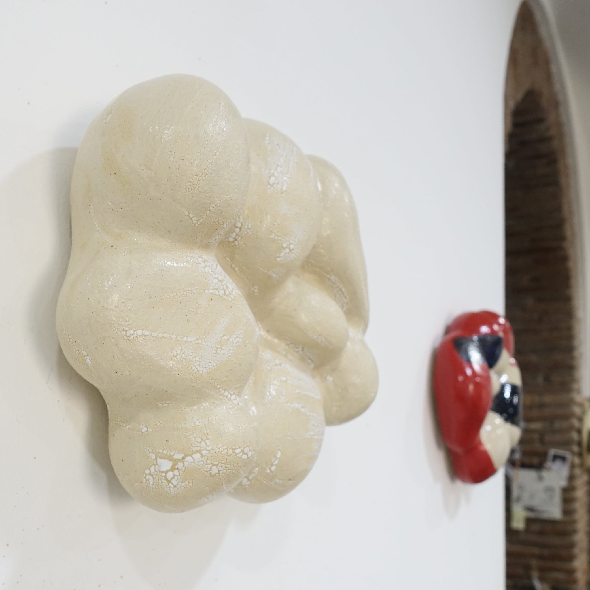 Seitenansicht einer keramischen Wandskulptur in organischer, wolkenartiger Form; helle, leicht cremeweiße Oberfläche mit feinen Rissstrukturen und weicher, gewölbter Modellierung. Im Hintergrund die Nuvola Rossa mit hellem Grundton, roten Flächen und abstrakten schwarzen und blauen Mustern.