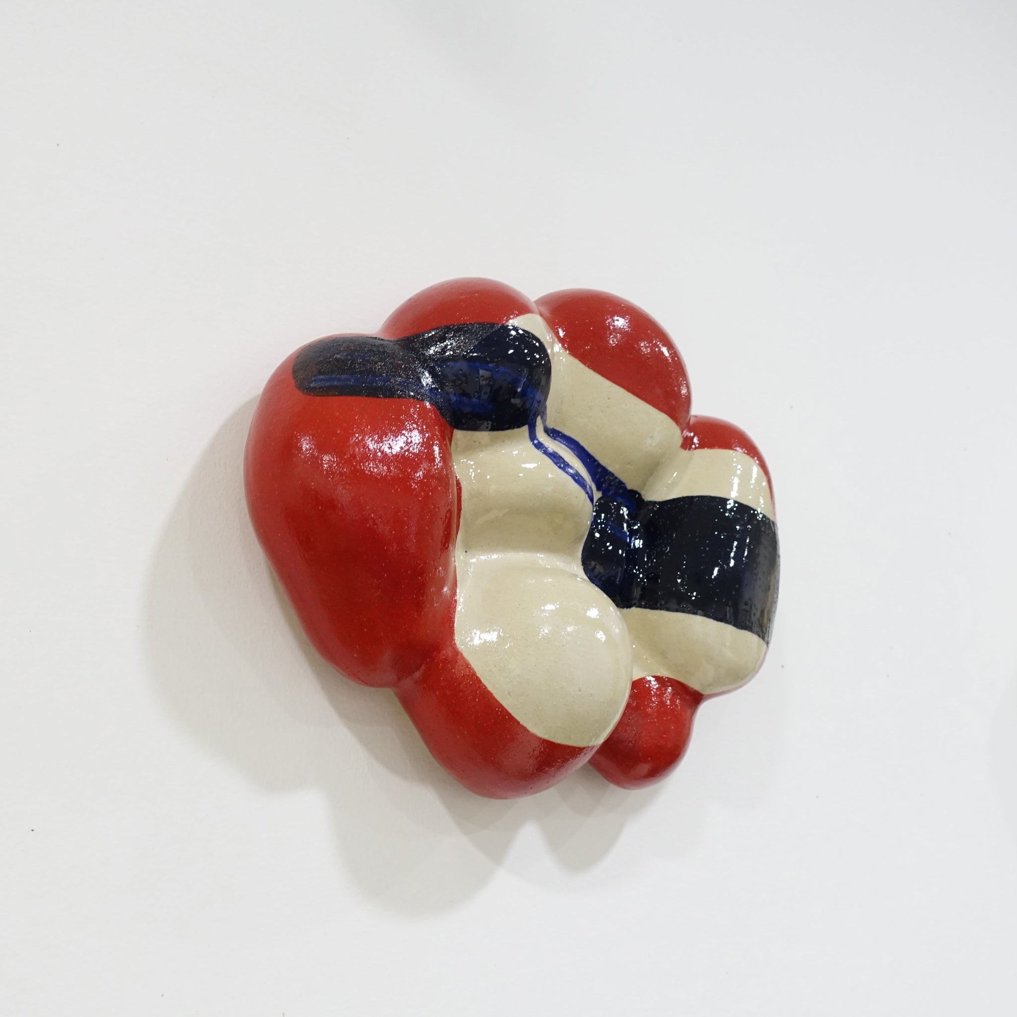 Leichte Seitenansicht einer keramischen Wandskulptur mit organisch gewölbter Form; cremefarbener Grundton mit leuchtend roten Flächen sowie dunkelblauen, gestischen Linien- und Flächenakzenten auf der Oberfläche.