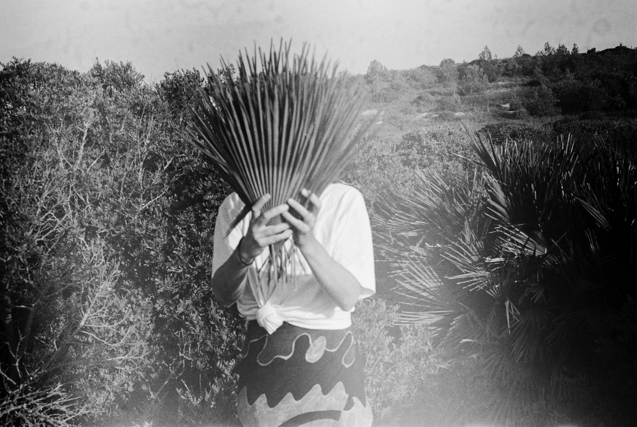 Schwarz-weiß Fotografie von Ysaline Ophoff, die gebündelte Palmblätter vor dem Gesicht hält; im Hintergrund die mediterrane Vegetation der Algarve im Süden Portugals.